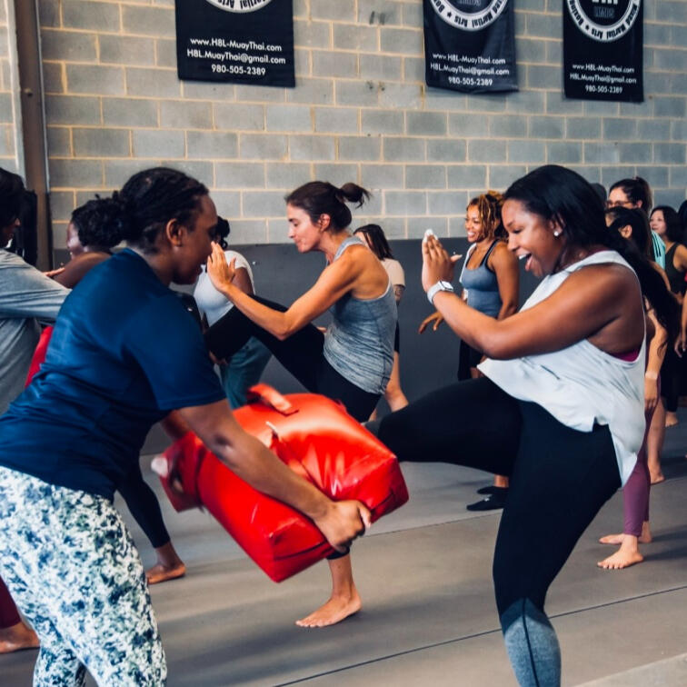 Multiple women taking part in a self-defense class. The woman on the right is wearing a grey tank, black leggings, and is kicking a red tombstone pad. The women in the background of the image are performing the same kicking technique.