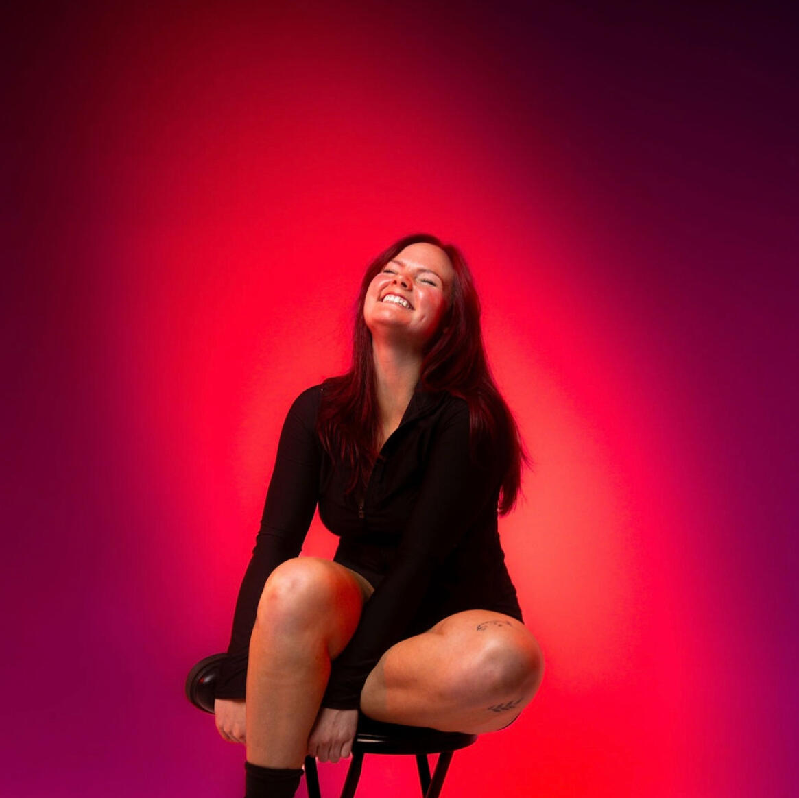 A 20-something, red-haired woman wearing a black top while sitting on a stool with one leg tucked under the other. Her eyes are closed while she tilts her head up and smiles big.