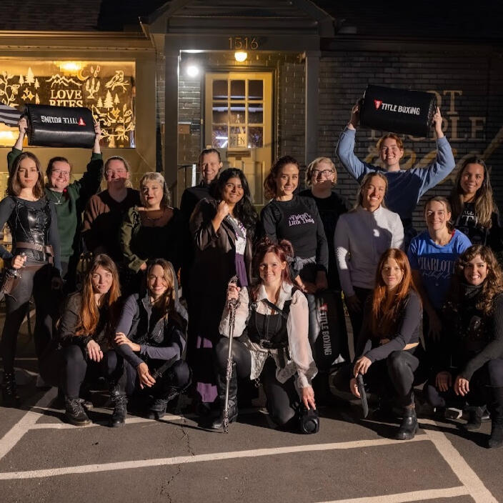 More than one dozen women dressed as fantasy characters and posing with swords, daggers, and tombstone hitting pads in front of a grey, brick building.
