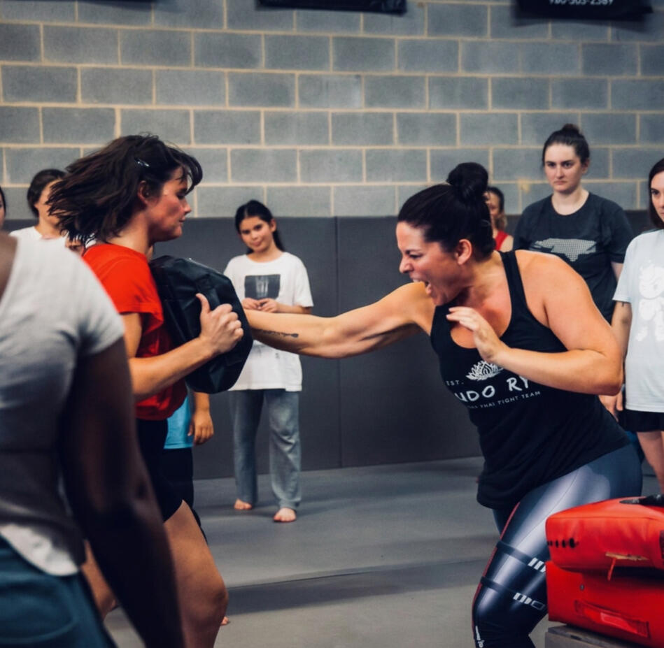 Multiple women taking part in a self-defense class. The woman on the right is hitting a tombstone pad with her right palm. Her mouth is open in a fighter's yell.